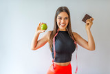 Attractive woman in sports clothes holding apple and chocolate bar isolated on grey. Dieting and eating habits concepts. Young cheerful girl in sports clothing holding chocolate and apple in hands.