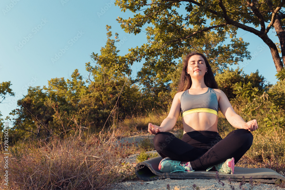 Young beautiful woman smiling and meditating in the Park. Sunset light. The concept of yoga, sports and meditation