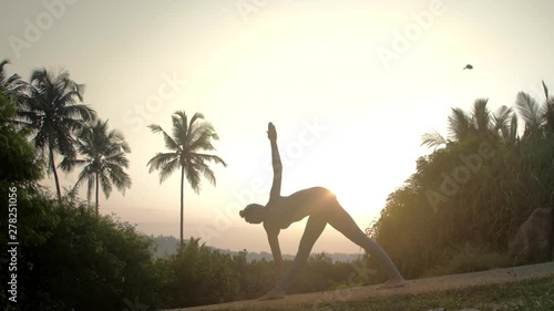 silhouette of woman standing in parivrtta trikonasana pose on track in tropical forest at sunrise light low angle shot slow motion. Concept fitness yoga wellness lifestyle photo