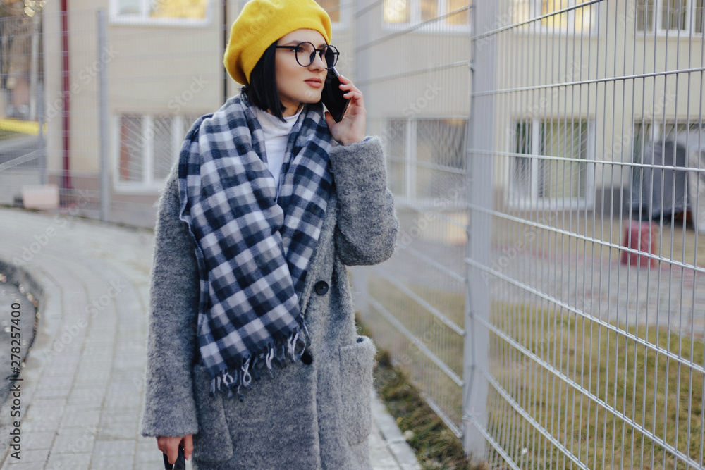 Fototapeta premium An attractive young girl wearing sunglasses in a coat walking down the street and talking on the phone and smiles