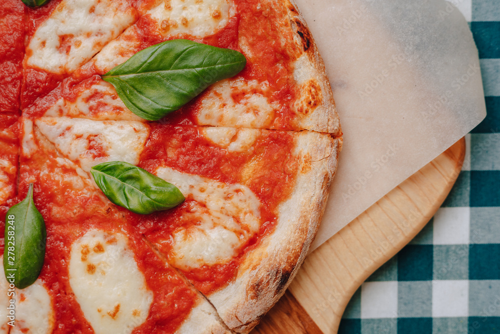 Neapolitan pizza with ham, cheese, arugula, basil, tomatoes sprinkled with cheese on a wooden board on a tablecloth in a cell with a place for text Neapolitan pizza with ham, cheese, arugula, basil, tomatoes sprinkled with cheese on a wooden board on a tablecloth in a cell with a place for text