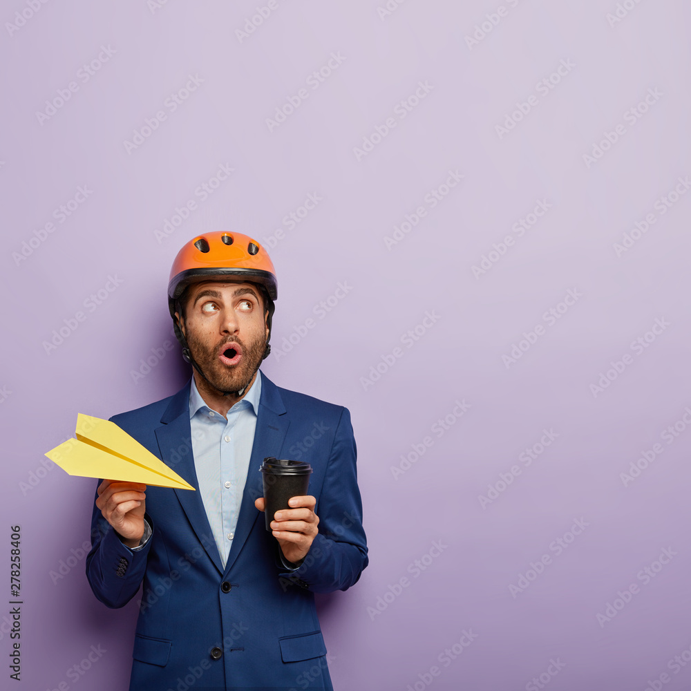 Industrial male engineer looks with stupefaction up, holds paper airplane, takeaway coffee, wears protective helmet and blue classy suit, has embarrased gaze, poses against purple background Industrial male engineer looks with stupefaction up, holds paper airplane, takeaway coffee, wears protective helmet and blue classy suit, has embarrased gaze, poses against purple background