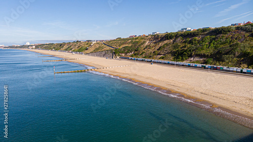 An aerial view of a majestic sandy beach with crystal blue water sea, groynes (breakwaters) and beach huts along a beautiful cliff with green vegetation under a blue and sunny sky