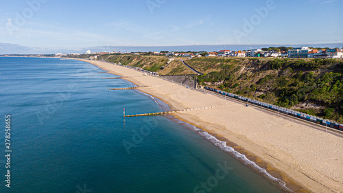 An aerial view of a majestic sandy beach with crystal blue water sea, groynes (breakwaters) and beach huts along a beautiful cliff with green vegetation under a blue and sunny sky