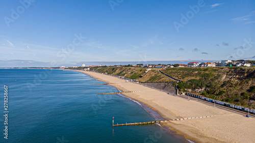 An aerial view of a majestic sandy beach with crystal blue water sea, groynes (breakwaters) and beach huts along a beautiful cliff with green vegetation under a blue and sunny sky