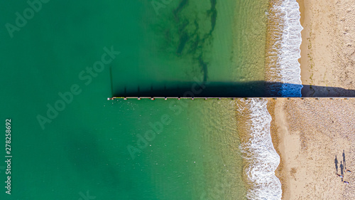 An aerial view of a groyne (breakwater) dividing a majestic cystal clear green water sea with crashing waves on a sunny sandy beach and three people running with their shade