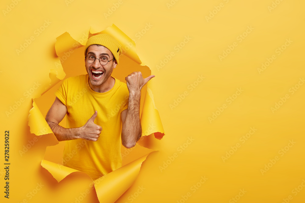 Smiling delighted guy points away, points with thumb and shows okay gesture, indicates at direction, wears yellow hat, t shirt, poses in ripped paper hole, has positive expression. Promotion concept - obrazy, fototapety, plakaty 