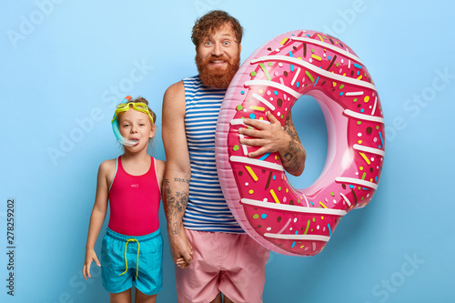 Indoor shot of delighted caring father in striped vest and shorts, holds inflated swimring, touches hand of little daughter in snorkel mask, have fun in water park, enjoy bathing, stand over blue wall photo