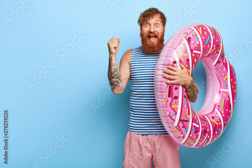 Yeah, finally summer! Joyful foxy holidaymaker clenches fist, rejoices coming holiday, dressed in casual outfit, carries inflated donut ring, shouts from pleasure, wins ticket for tour abroad