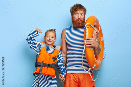 Happy small child in orange lifejacket raises arm and shows muscle, hold hands with father. Red haired dad in sailor vest and short, carries lifebuoy for safe swimming together with daughter.