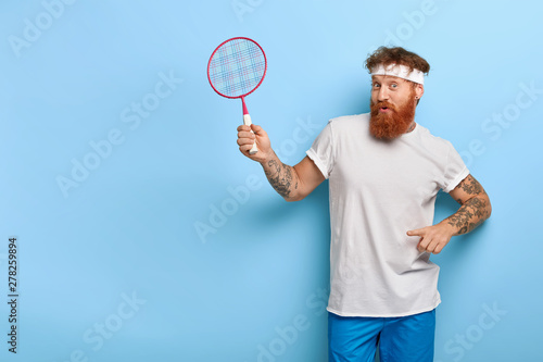 Sport is best motivation. Sporty athletic redhead man poses with tennis racket, points at blank space, wears white headband, t shirt and blue shorts, invites to play with him, happy win game © wayhome.studio 