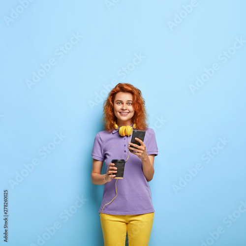 Smiling tender red head woman in colorful clothing, stands with smart phone and takeaway coffee, chats online, checks newsfeed, connected to wireless internet. Happy female student with gadget