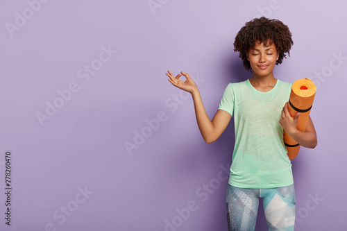 Young relaxed dark skinned woman practices yoga, meditates indoor for feeling relaxation, has eyes closed, holds fitness mat, dressed in active wear, isolated on purple background blank space on left