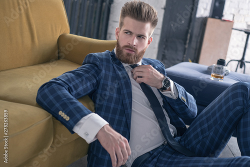 Impressive look. Handsome young man in full suit looking over his shoulder while sitting on the floor at home