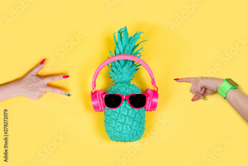 Pineapple in pink headphones and glasses and woman hаnds on a yellow background