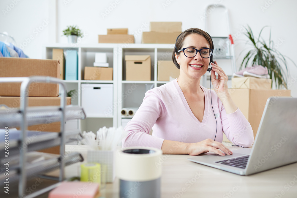 Smiling young helpdesk operator in headset talking in microphone while sitting in front of laptop by desk Smiling young helpdesk operator in headset talking in microphone while sitting in front of laptop by desk