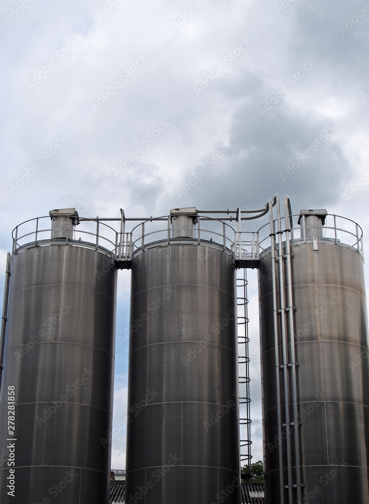 a group of three tall steel chemical storage tanks with connecting pipes and ladders against a blue cloudy sky