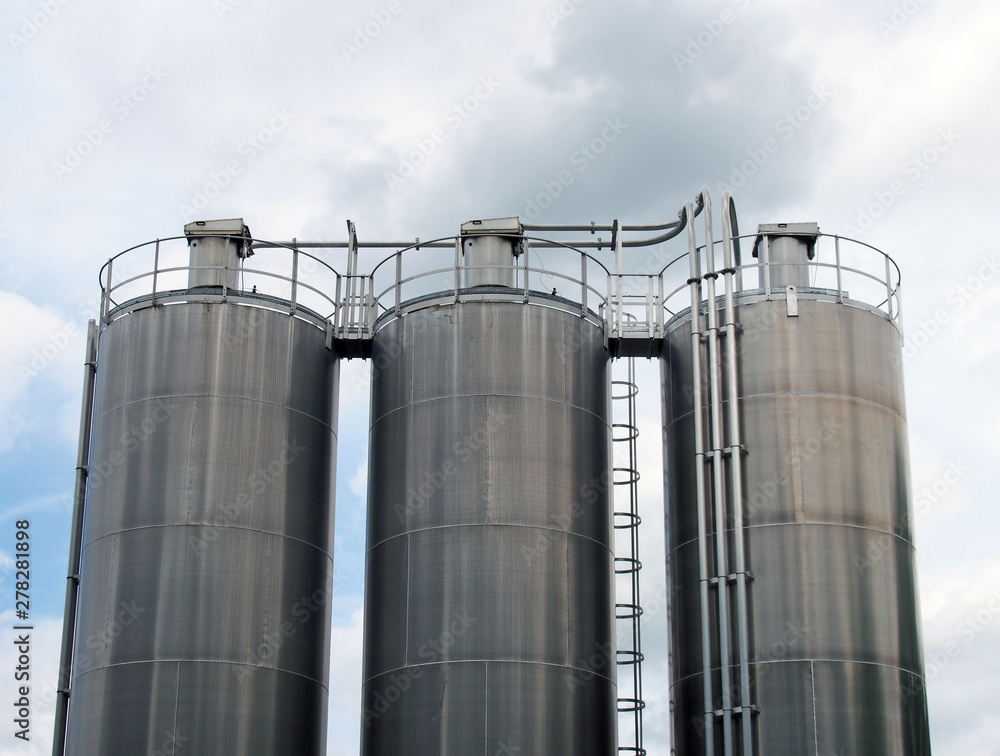 a group of three tall steel chemical storage tanks with connecting pipes and ladders against a blue cloudy sky a group of three tall steel chemical storage tanks with connecting pipes and ladders against a blue cloudy sky