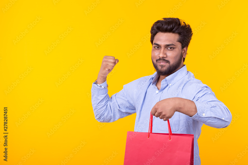 Young handsome indian man with a beard is holding red shopping bags posing on a yellow background with copy space. Supermarket shopping concept. Young handsome indian man with a beard is holding red shopping bags posing on a yellow background with copy space. Supermarket shopping concept.