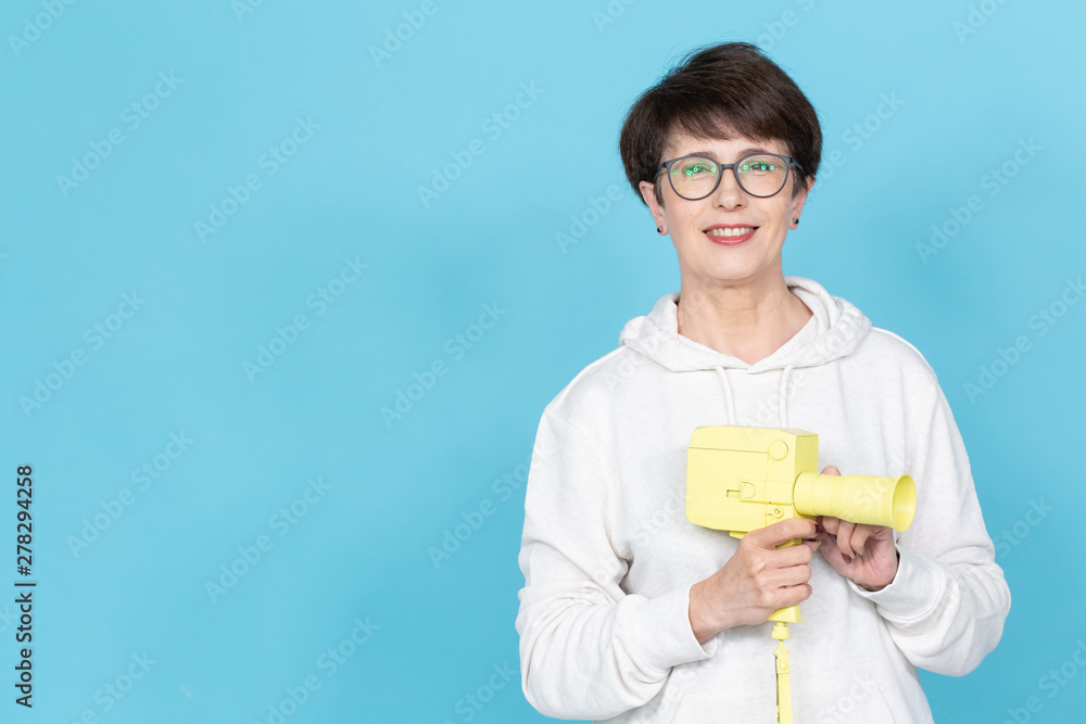 Middle-aged woman with a short haircut in a sweater and glasses holding a yellow vintage movie camera on blue background with copy space. Concept of video shooting Middle-aged woman with a short haircut in a sweater and glasses holding a yellow vintage movie camera on blue background with copy space. Concept of video shooting