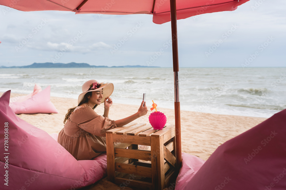 On vacation at Pattaya beach, asian happy young beautiful woman in dress cloth video call with friends on a pink chair in luxury cafe.