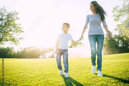 Happy attractive mother walks with her little cute son and holding his hand on sunset in summer green park