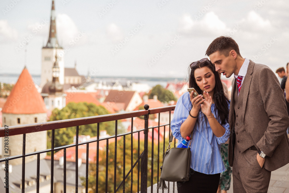 The young couple standing against the background of the view on the city and looking at the phone