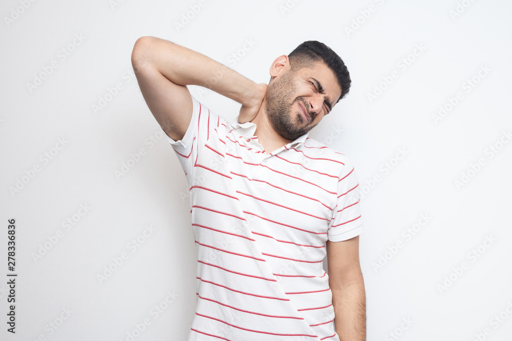 Neck ache or pain. Portrait of handsome bearded young man in striped t-shirt standing and holding his painful neck. indoor studio shot, isolated on white background. Neck ache or pain. Portrait of handsome bearded young man in striped t-shirt standing and holding his painful neck. indoor studio shot, isolated on white background.