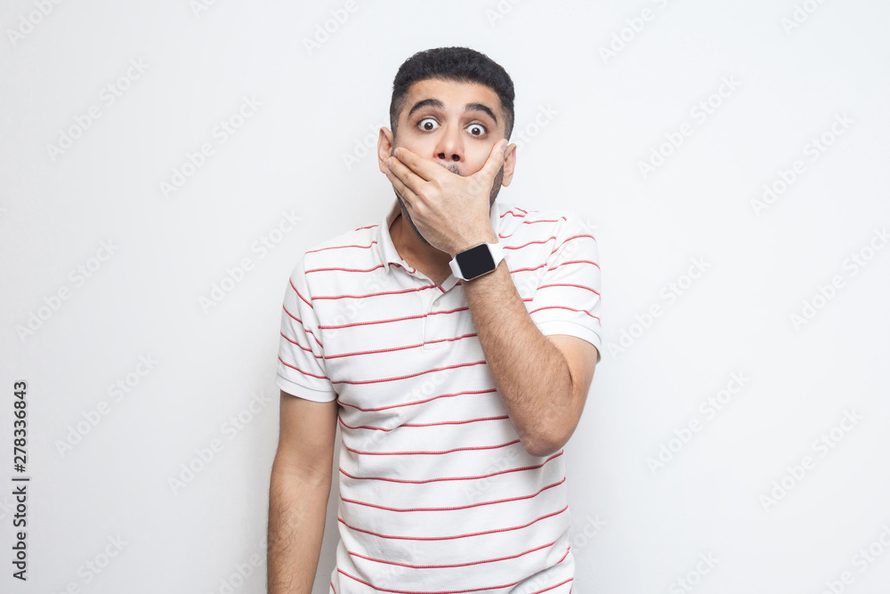 i will be quiet. Portrait of shocked bearded young man in striped t-shirt standing closing his mouth with hands and looking at camera with big eyes. indoor studio shot, isolated on white background. i will be quiet. Portrait of shocked bearded young man in striped t-shirt standing closing his mouth with hands and looking at camera with big eyes. indoor studio shot, isolated on white background.