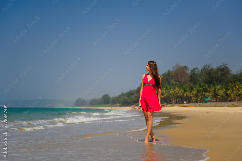 Young slender long-haired brunette in red dress walks barefoot on tropical beach along the surf against the sea and blue sky on a hot day. In the distance a green hill and people on the shore. Young slender long-haired brunette in red dress walks barefoot on tropical beach along the surf against the sea and blue sky on a hot day. In the distance a green hill and people on the shore.