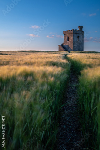 A portrait shot of a lookout fort standing proud in a field of wheat at sunset, path in the field leading to the fort. photo