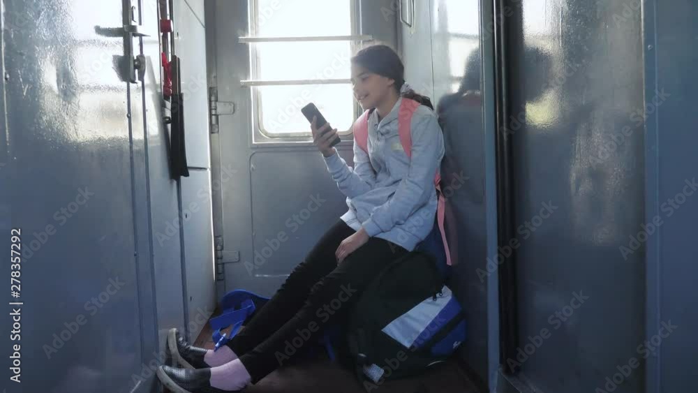 Teenager girl traveler with a backpack sitting on bags backpacks at the window of the car with a smartphone. Travel rail carriage concept. the girl in the train at the window corresponds to the girl Teenager girl traveler with a backpack sitting on bags backpacks at the window of the car with a smartphone. Travel rail carriage concept. the girl in the train at the window corresponds to the girl