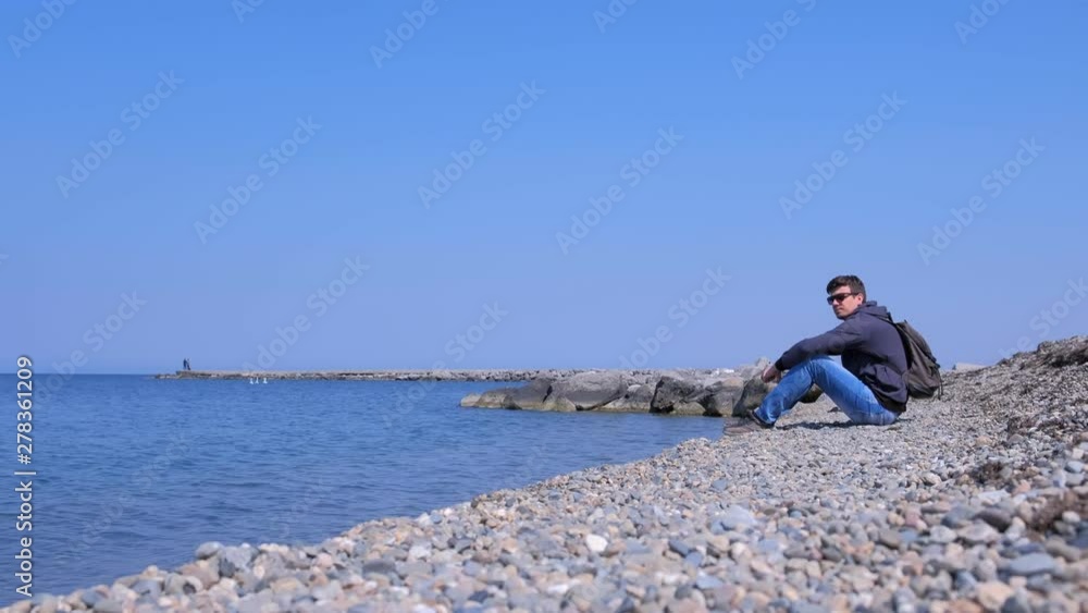 Young man traveler sit on stone beach and looks at sea admires seascape on spring vacation, side view. He is with backpack in sunglasses, sweatshirt and jeans. Travel concept. Young man traveler sit on stone beach and looks at sea admires seascape on spring vacation, side view. He is with backpack in sunglasses, sweatshirt and jeans. Travel concept.