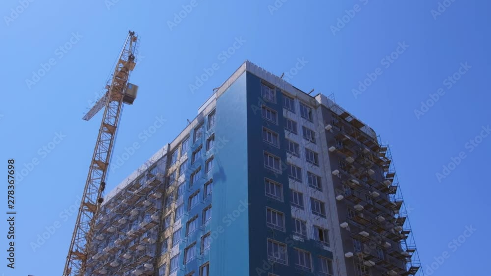 Multi-storey house under construction on the background of the blue sky next to the construction crane. High rise building in the city in new modern district. Multi-storey house under construction on the background of the blue sky next to the construction crane. High rise building in the city in new modern district.