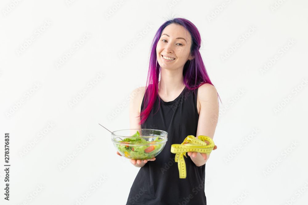Slim young asian girl with colored hair is holds a measuring tape and holding a light vegetable salad in her hands posing on a white background. Healthy eating concept. Copyspace. Slim young asian girl with colored hair is holds a measuring tape and holding a light vegetable salad in her hands posing on a white background. Healthy eating concept. Copyspace.