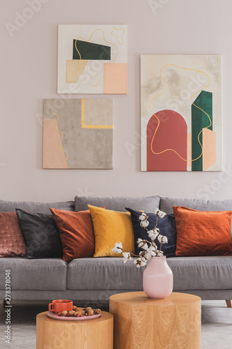 Yellow, orange, black and brown pillows on comfortable grey scandinavian sofa in bright living room interior with abstract paintings on the wall