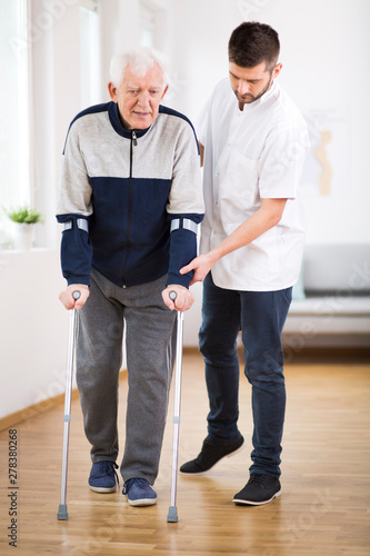 Elderly man walking on crutches and a helpful male nurse supporting him © Photographee.eu