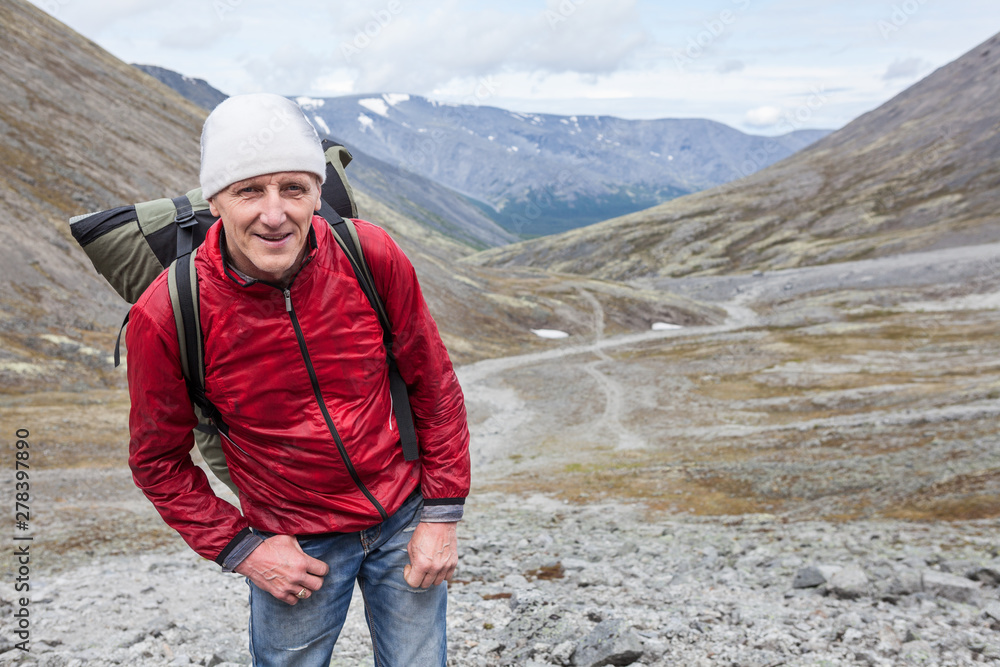 Happy and smiling mature backpacker looking at camera while hiking in mountains, copy space