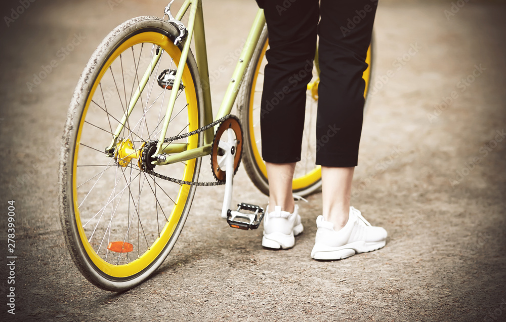 Fototapeta premium A girl in black pants and white sneakers stands near her beautiful bike is yellow-green in the Park in the summer.