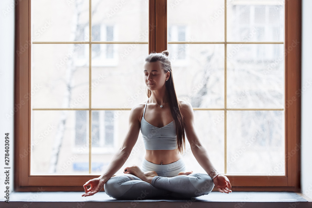 Young beautiful girl meditates in lotus pose sitting by the large windows of a country house. Concept of relaxation and meditation. Lotus exercise, Ardha Padmasana pose, mudra gesture, silhouette