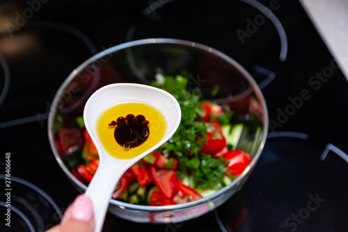 Preparation of salad from fresh vegetables, cucumbers, tomatoes and greens with a white plastic spoon containing a salad dressing made from olive oil, balsamic vinegar and soy bean sauce photo