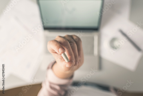 Top view. Tired business woman holding a headache pill on the workplace with glass of water on the background of graphics and charts printed on the paper.