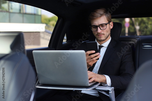 Handsome confident man in full suit looking at his smart phone while sitting in the car and using laptop. © ty