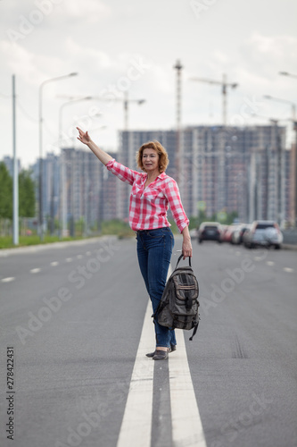Middle age Caucasian woman waving hand for stop the car, standing on the median strip line of urban road