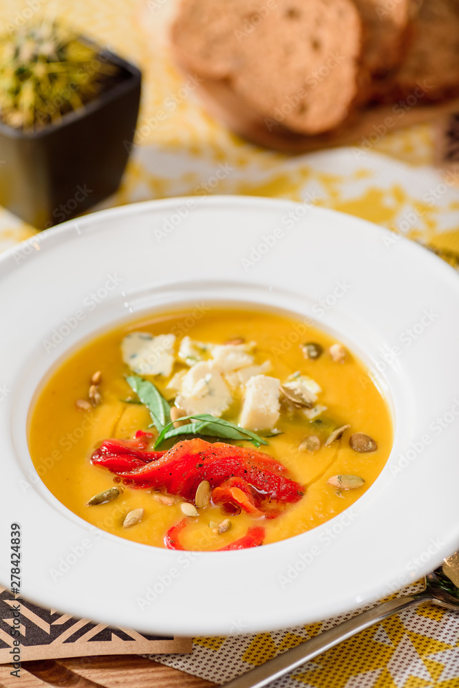 pumpkin cream soup with cheese Dor Blue, bell pepper, pumpkin seeds in a white plate on yellow set on a wooden table in a restaurant. Pumpkin soup with toast on a table in a restaurant. close-up pumpkin cream soup with cheese Dor Blue, bell pepper, pumpkin seeds in a white plate on yellow set on a wooden table in a restaurant. Pumpkin soup with toast on a table in a restaurant. close-up