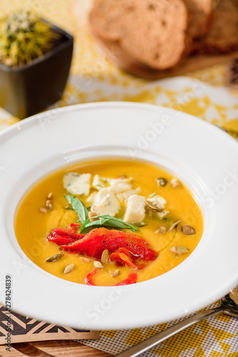 pumpkin cream soup with cheese Dor Blue, bell pepper, pumpkin seeds in a white plate on yellow set on a wooden table in a restaurant. Pumpkin soup with toast on a table in a restaurant. close-up