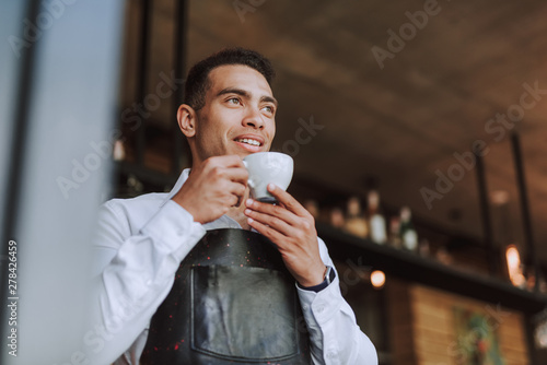 Handsome young man in black apron drinking coffee in cafe