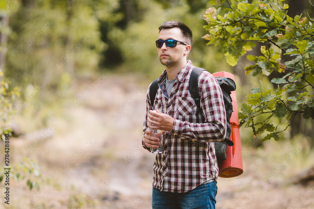 Fototapeta premium Hiker Drinking Water In Forest. Tired man drinking water from bottle in woodland. Handsome Traveler With Backpack And Flask In Forest. vacation, outdoor lifestyle freedom concept. Adventures hiking
