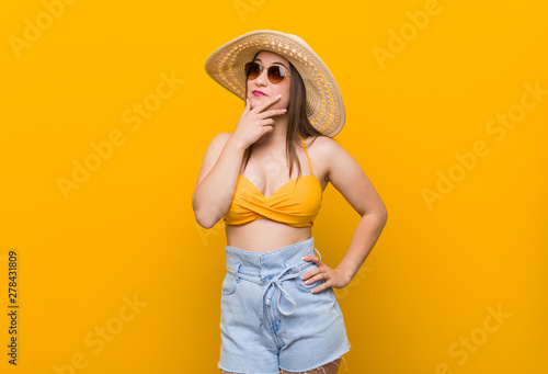 Young caucasian woman wearing a straw hat, summer look looking sideways with doubtful and skeptical expression.