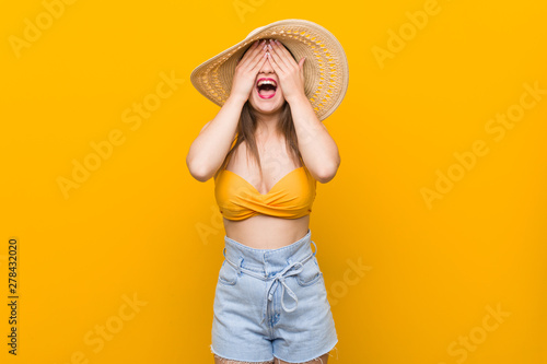 Young caucasian woman wearing a straw hat, summer look covers eyes with hands, smiles broadly waiting for a surprise.